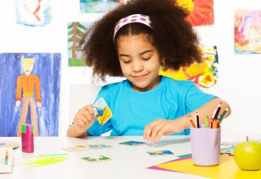 African girl putting matching cards during developmental game on table while sitting in playroom with wall behind full of children drawings